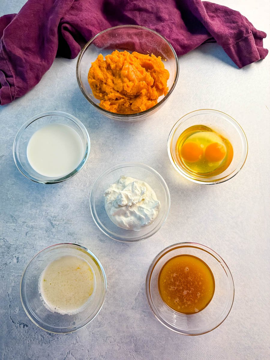 A lineup of wet ingredients in small glass bowls including mashed sweet potatoes, milk, eggs, sour cream, melted butter, and honey ready to be mixed.