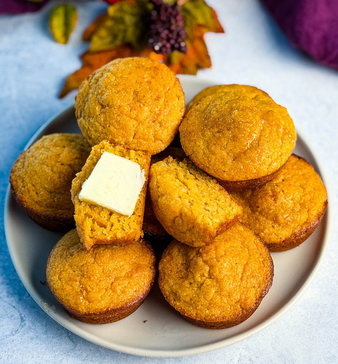 A plate of golden-brown Jiffy sweet potato cornbread muffins stacked high, with one muffin split open to show the moist texture and topped with a pat of butter.