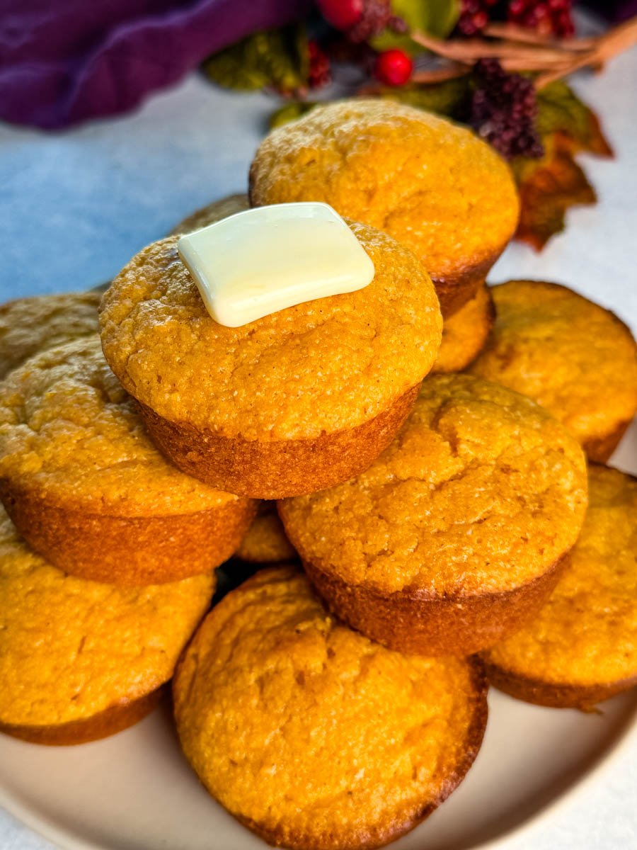 A close-up of warm sweet potato cornbread muffins stacked on a plate, with a melting square of butter on top.