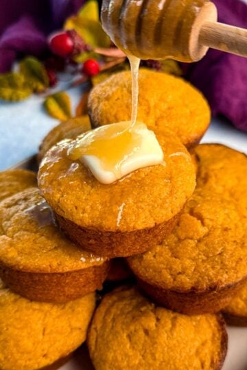 A stack of golden-brown Jiffy sweet potato cornbread muffins on a plate, topped with melting butter and drizzled with honey for a glossy, irresistible finish.