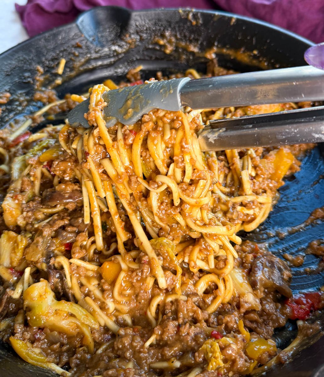 Tongs lifting glossy Mongolian ground beef noodles out of a skillet, showing the thick, flavorful sauce coating every strand.