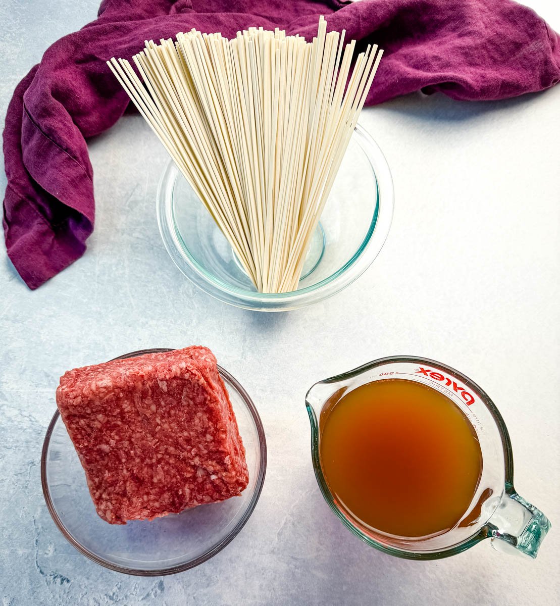 Overhead shot of three main ingredients for one pot Mongolian ground beef with udon noodles—raw ground beef, uncooked udon noodles in a glass bowl, and a measuring cup of broth. A purple kitchen towel sits in the background on a light surface.