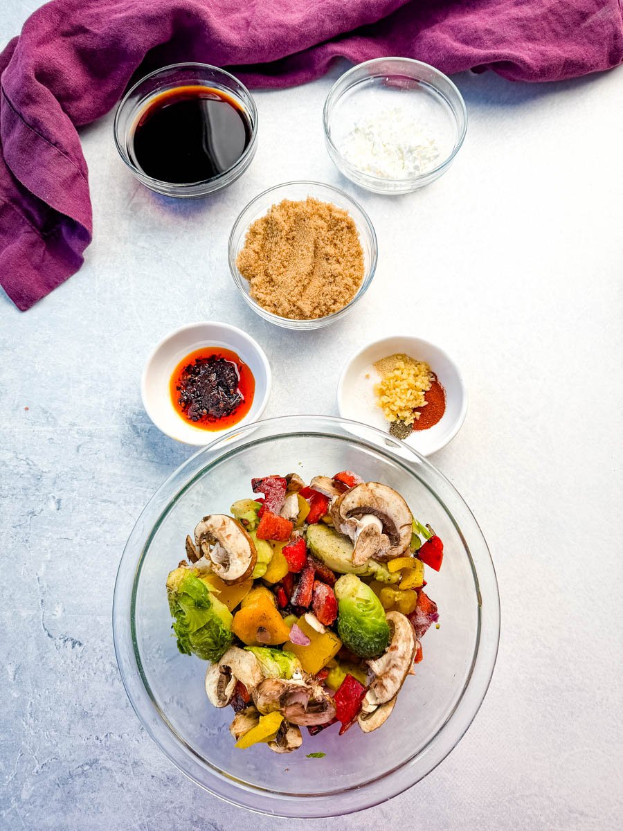 Overhead photo showing prepped ingredients for Mongolian ground beef with udon noodles, including a bowl of chopped mixed vegetables (Brussels sprouts, bell peppers, mushrooms), and small bowls of soy sauce, brown sugar, garlic, cornstarch, and seasonings arranged neatly on a light countertop with a purple cloth in the background.