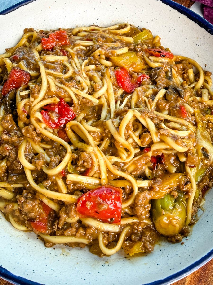 Close-up shot of Mongolian ground beef with udon noodles served in a white bowl with a blue rim. The noodles are coated in a rich brown sauce with red and yellow bell peppers mixed in.