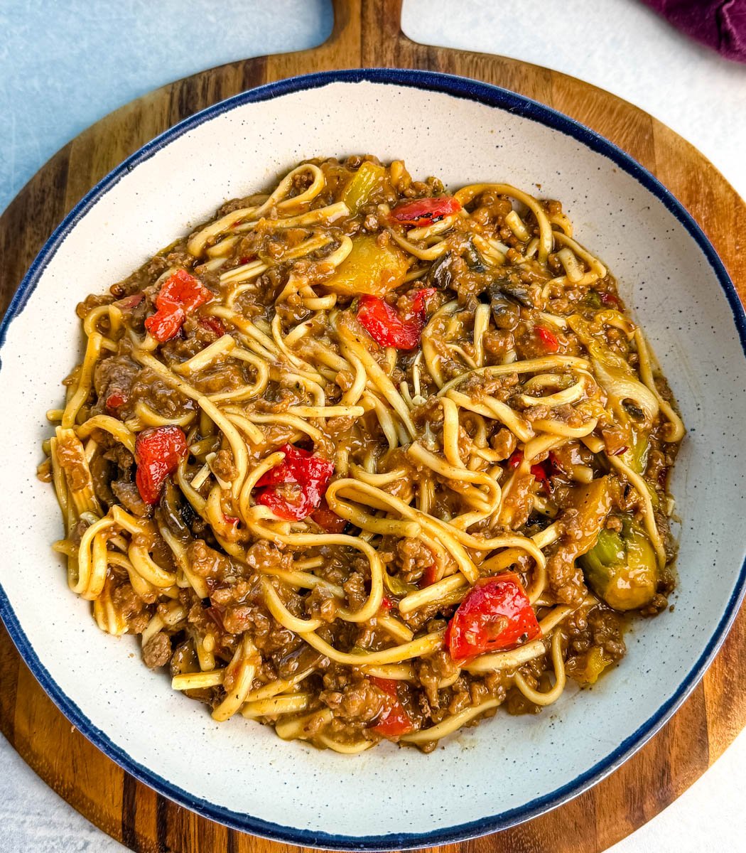 Overhead view of Mongolian ground beef and noodles in a white bowl on a wooden board. The dish looks saucy and hearty, with colorful vegetables and thick udon noodles.