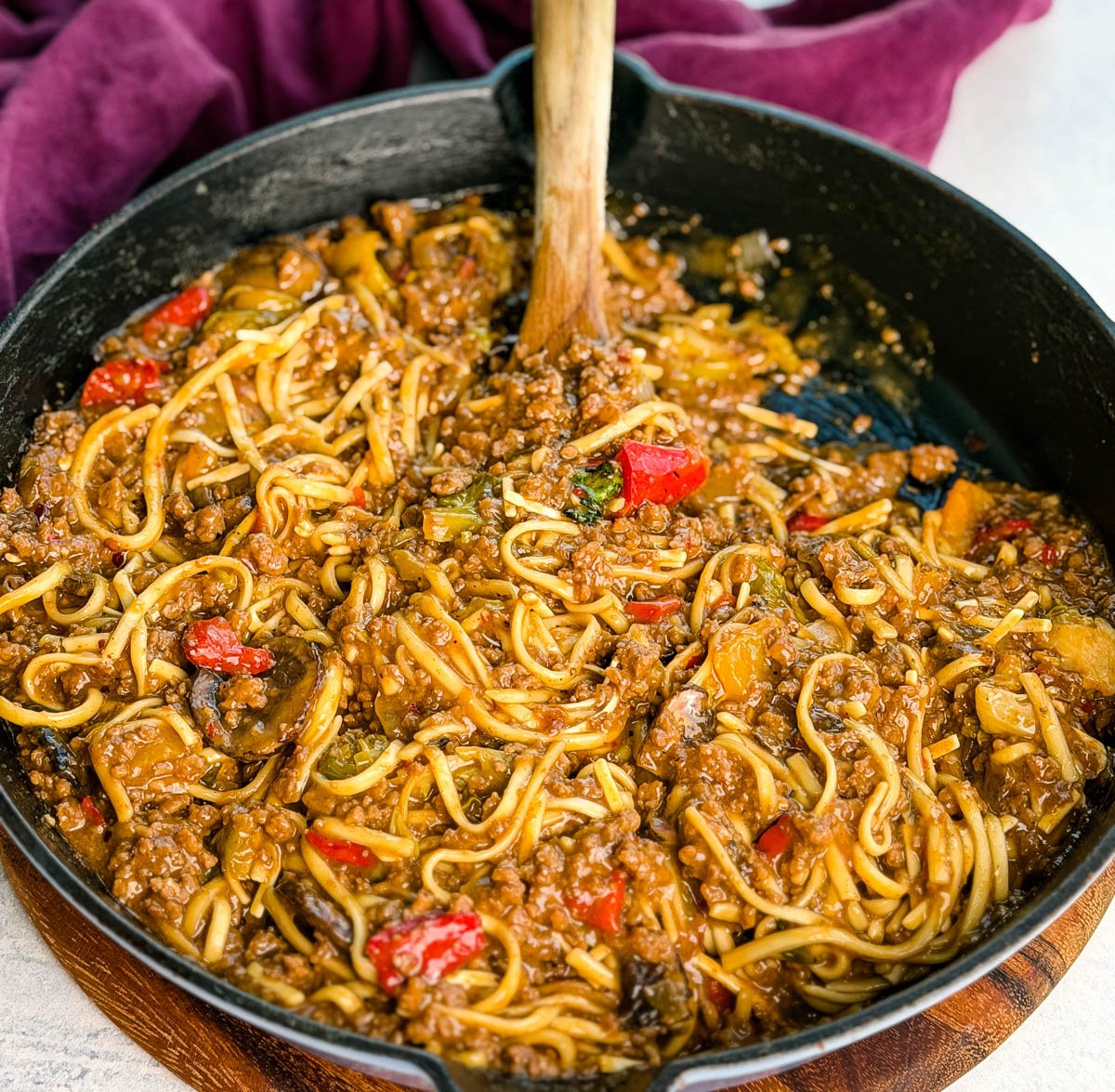 Overhead photo of the entire skillet filled with Mongolian ground beef noodles, wooden spoon in the pan, and a purple napkin in the background. The glossy sauce and colorful veggies stand out against the black skillet.