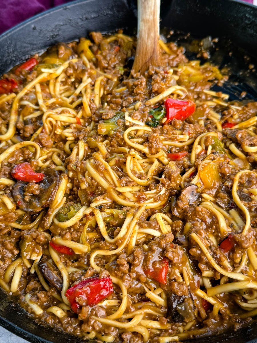 Close-up shot showing saucy Mongolian ground beef noodles with red peppers, mushrooms, and bits of green vegetables, highlighting the glossy, caramelized texture of the sauce.