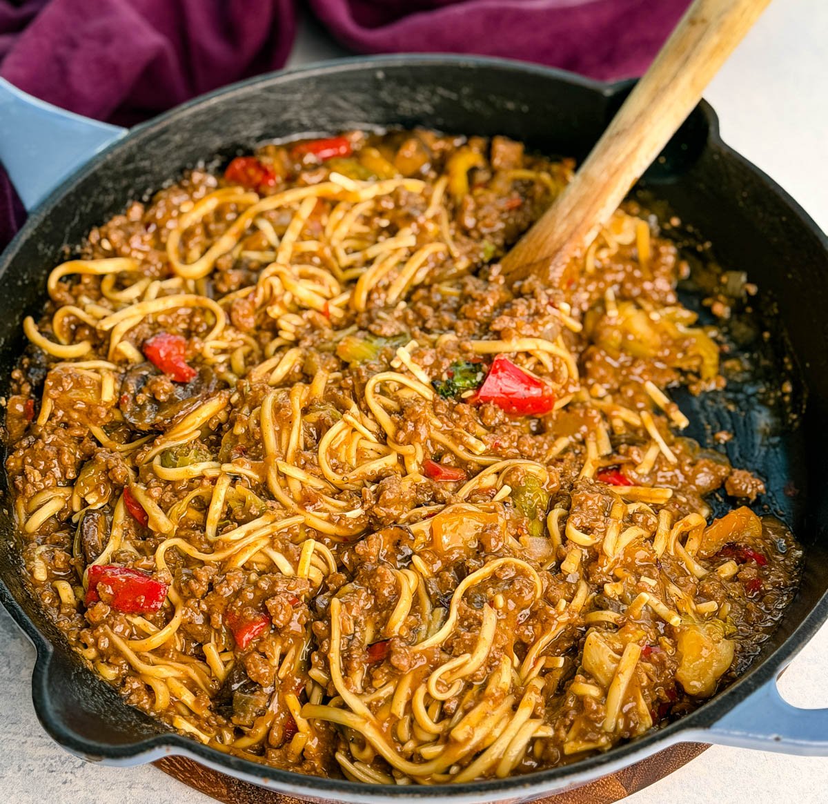 Overhead photo of the entire skillet filled with Mongolian ground beef noodles, wooden spoon in the pan, and a purple napkin in the background. The glossy sauce and colorful veggies stand out against the black skillet.