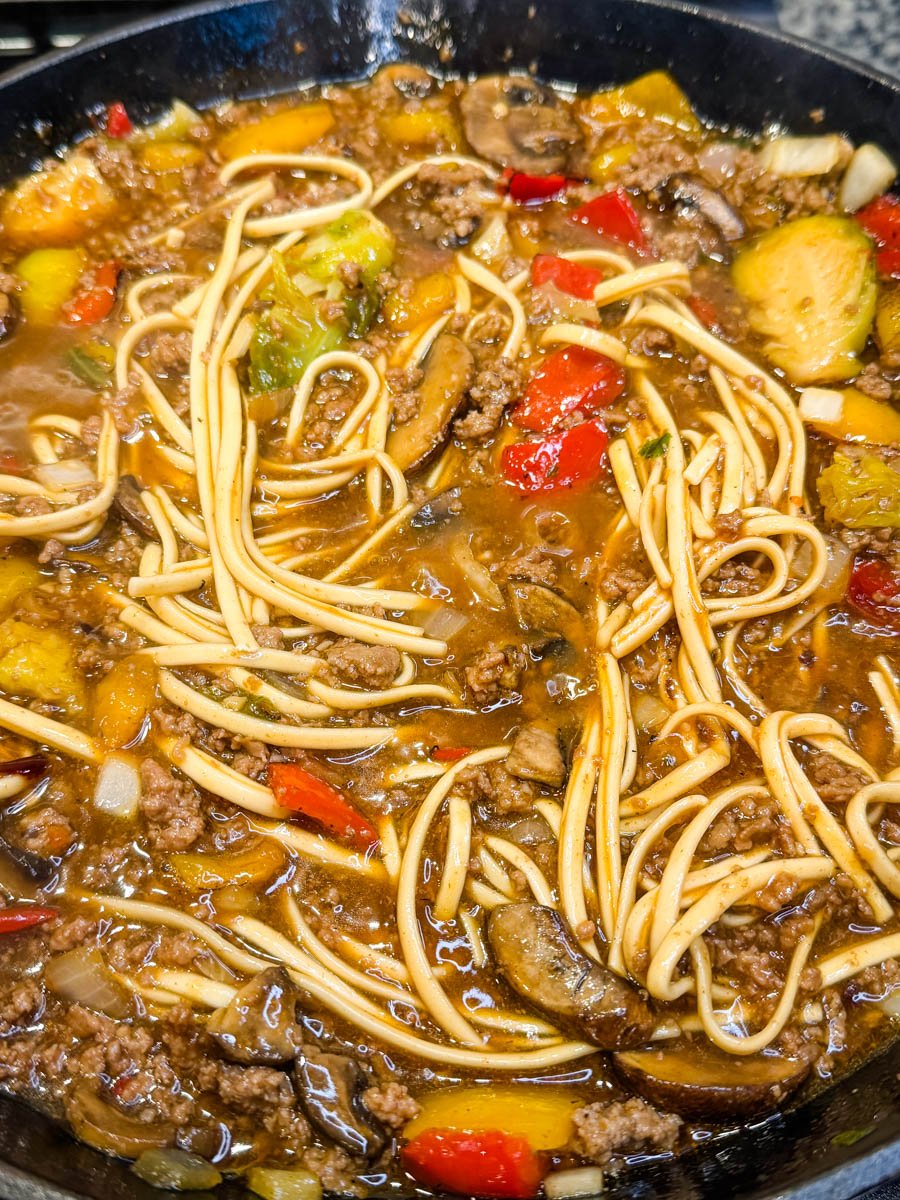 Close-up shot showing saucy Mongolian ground beef noodles with red peppers, mushrooms, and bits of green vegetables, highlighting the glossy, caramelized texture of the sauce.