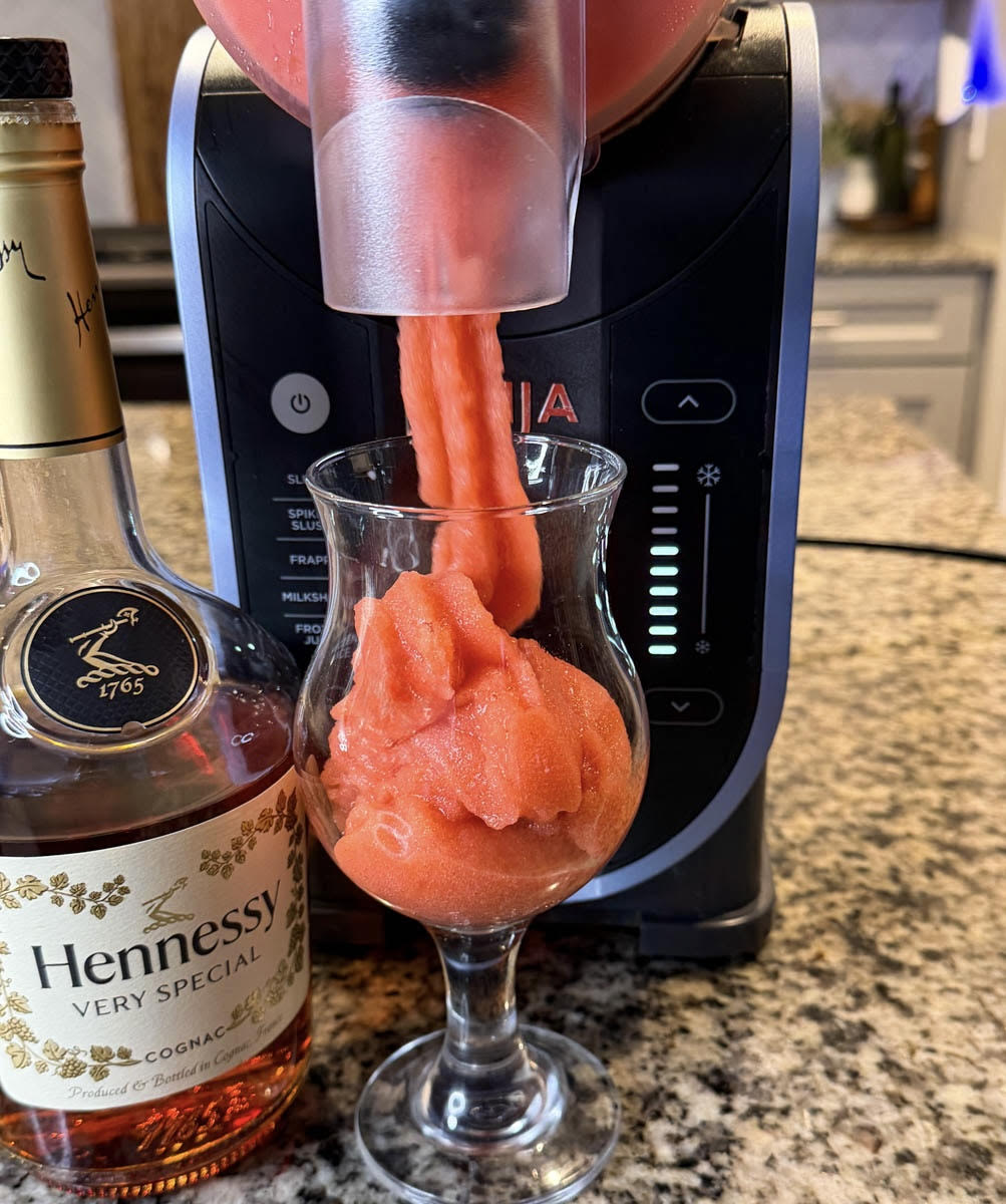 Ninja Slushi machine dispensing a smooth red Hennessy slushie into a glass beside a bottle of Hennessy on a kitchen counter.