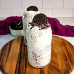 Two Oreo McFlurry-style Ninja Slushi drinks with crushed cookies and whole Oreos on top, placed on a wooden board in front of a bowl of Oreos and a purple cloth.