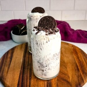 Two Oreo McFlurry-style Ninja Slushi drinks with crushed cookies and whole Oreos on top, placed on a wooden board in front of a bowl of Oreos and a purple cloth.