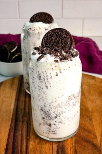 Two Oreo McFlurry-style Ninja Slushi drinks with crushed cookies and whole Oreos on top, placed on a wooden board in front of a bowl of Oreos and a purple cloth.