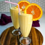 Two tall glasses filled with orange Julius slushies, each garnished with a large orange slice and a striped straw, placed on a wooden board with a hexagon-tile backsplash behind them.
