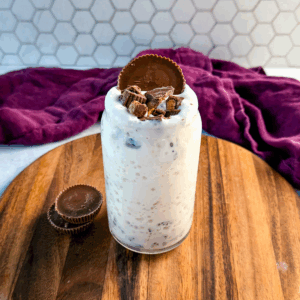 A full glass jar of peanut butter cup milkshake topped with chopped peanut butter cups and a whole peanut butter cup, sitting on a wooden board with two extra peanut butter cups beside it and a purple cloth in the background.