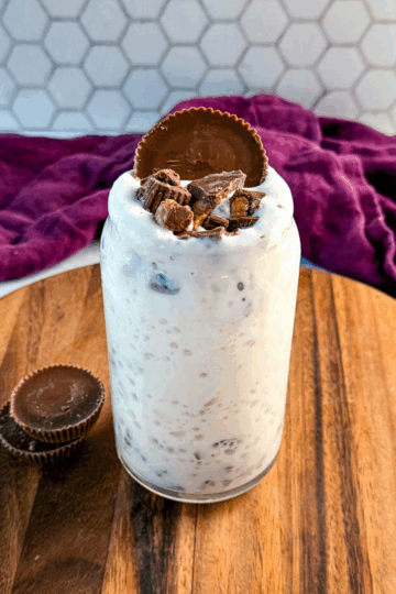 A full glass jar of peanut butter cup milkshake topped with chopped peanut butter cups and a whole peanut butter cup, sitting on a wooden board with two extra peanut butter cups beside it and a purple cloth in the background.