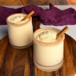 Two glasses of frozen spiked eggnog on a wooden board, each topped with a cinnamon stick and rimmed with cinnamon sugar, with a purple cloth in the background.