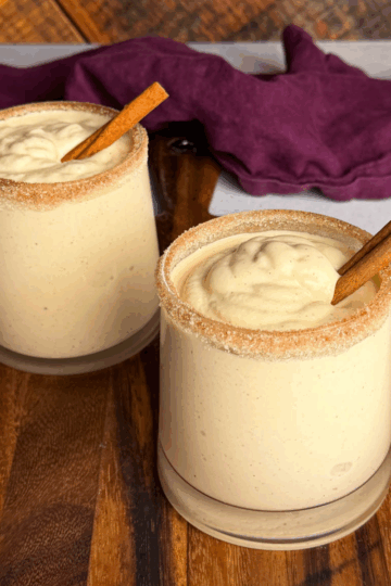Two glasses of frozen spiked eggnog on a wooden board, each topped with a cinnamon stick and rimmed with cinnamon sugar, with a purple cloth in the background.