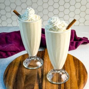 Two glasses of Ninja Slushi eggnog on a wooden board, garnished with whipped cream and cinnamon sticks, against a tiled kitchen wall.