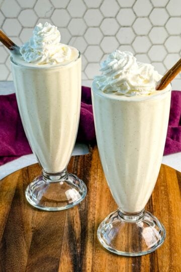 Two glasses of Ninja Slushi eggnog on a wooden board, garnished with whipped cream and cinnamon sticks, against a tiled kitchen wall.
