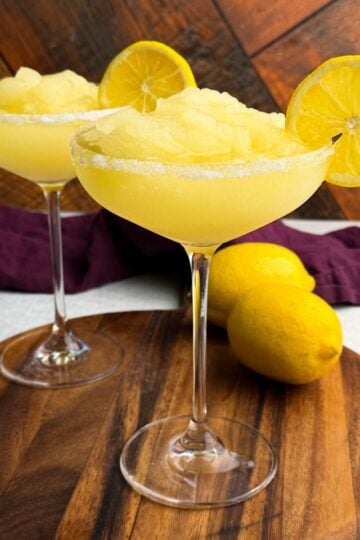 Two coupe glasses filled with pale yellow frozen lemon drop slush, rimmed with sugar and garnished with lemon slices, placed on a round wooden board with whole lemons and a purple cloth in the background.