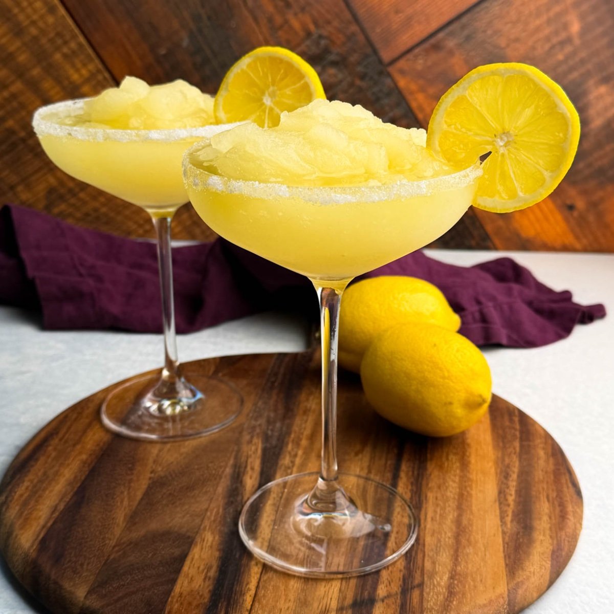 Two coupe glasses filled with pale yellow frozen lemon drop slush, rimmed with sugar and garnished with lemon slices, placed on a round wooden board with whole lemons and a purple cloth in the background.