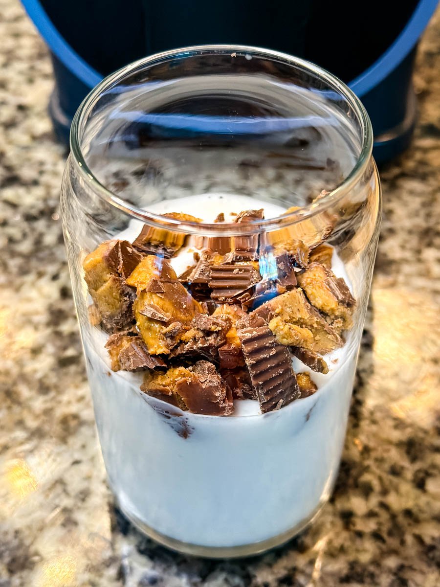 A glass filled with milk and chopped peanut butter cups before mixing, sitting on a granite countertop with the Ninja Slushi machine in the background.