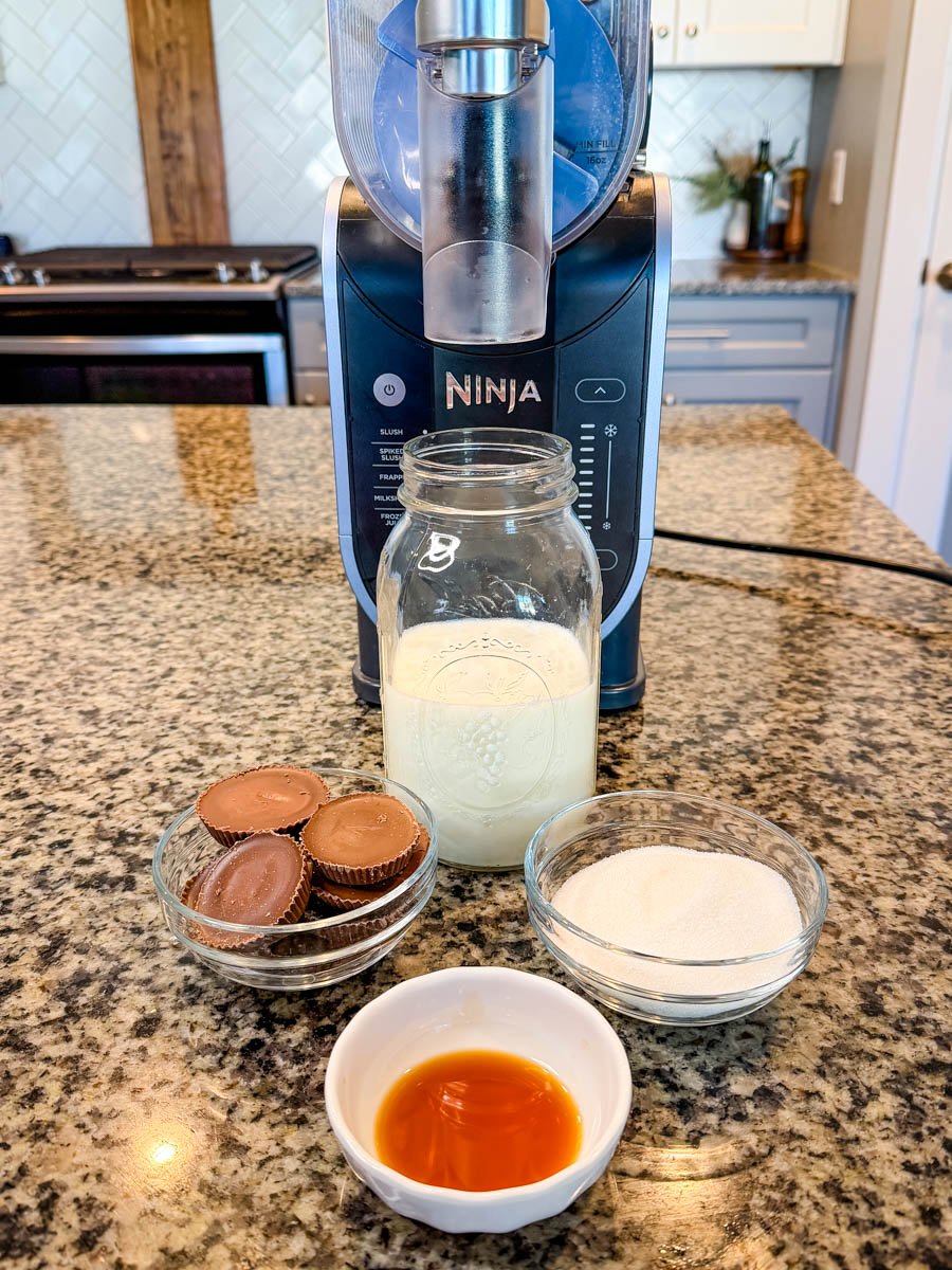 Ingredients for a peanut butter cup milkshake arranged in front of the Ninja Slushi machine: a jar of milk, a bowl of peanut butter cups, a bowl of sugar, and a small dish of vanilla extract.