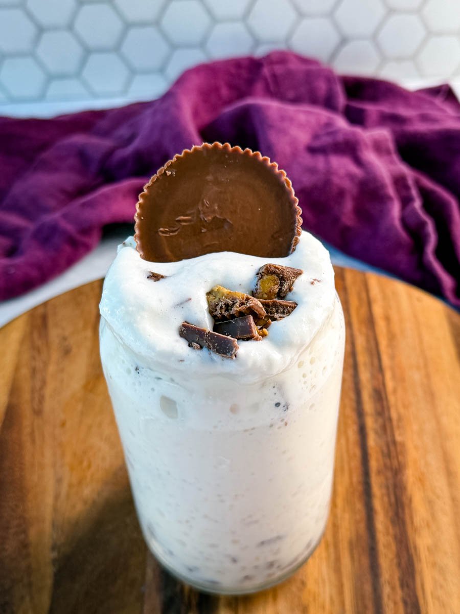 A close-up of a thick peanut butter cup milkshake in a glass jar, topped with crushed peanut butter cups and a whole peanut butter cup on top, set in front of a purple cloth on a wooden board.