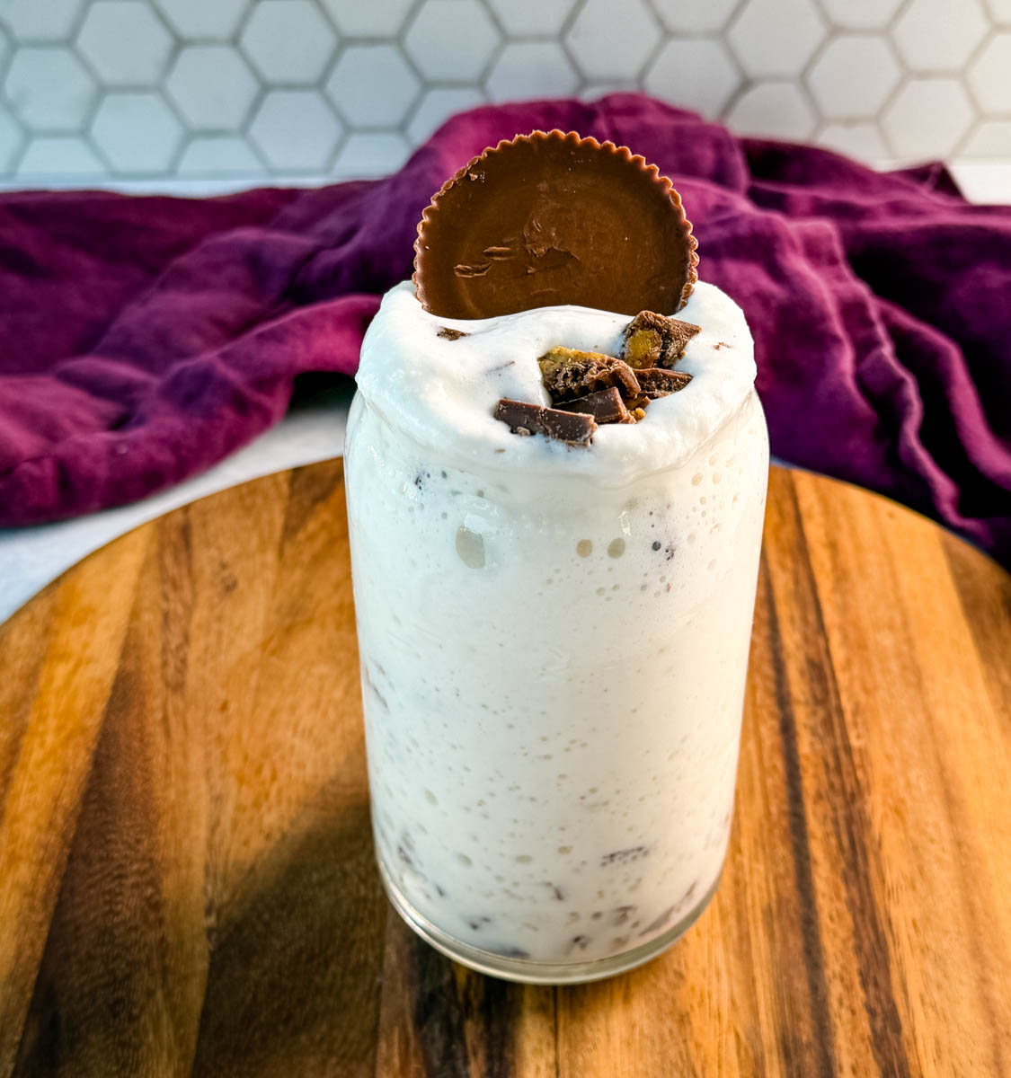 A creamy peanut butter cup milkshake in a glass jar, topped with chopped peanut butter cups and a whole peanut butter cup pressed into the top, with a purple cloth in the background.
