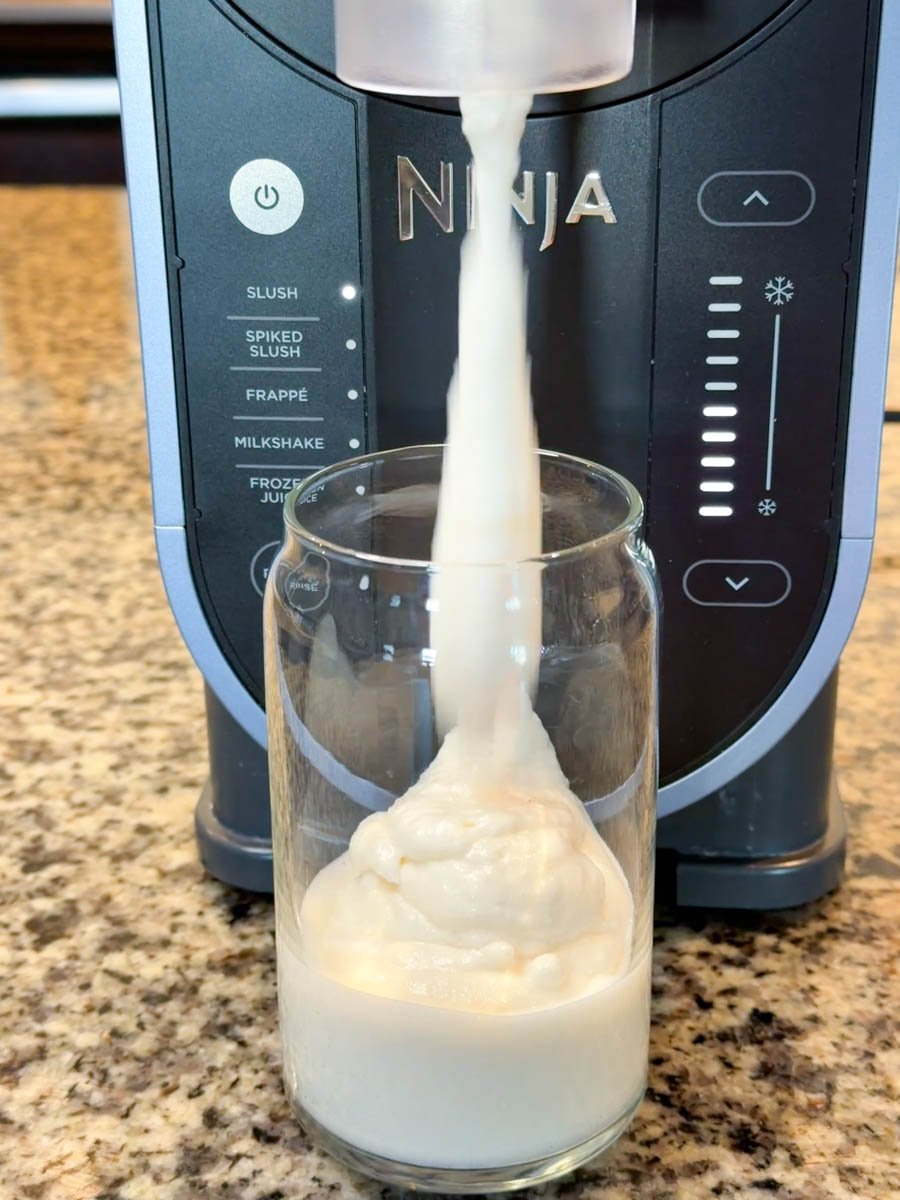 A creamy milkshake pouring out of the Ninja Slushi machine into a tall glass jar on a granite countertop, forming a thick swirl at the bottom.