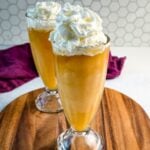 Two tall glasses filled with golden Butter Beer slushies, each topped with a large swirl of whipped cream, set on a wooden board with a hexagon-tile backsplash behind them.