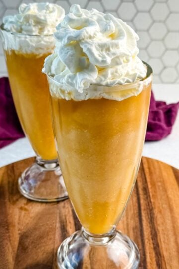 Two tall glasses filled with golden Butter Beer slushies, each topped with a large swirl of whipped cream, set on a wooden board with a hexagon-tile backsplash behind them.