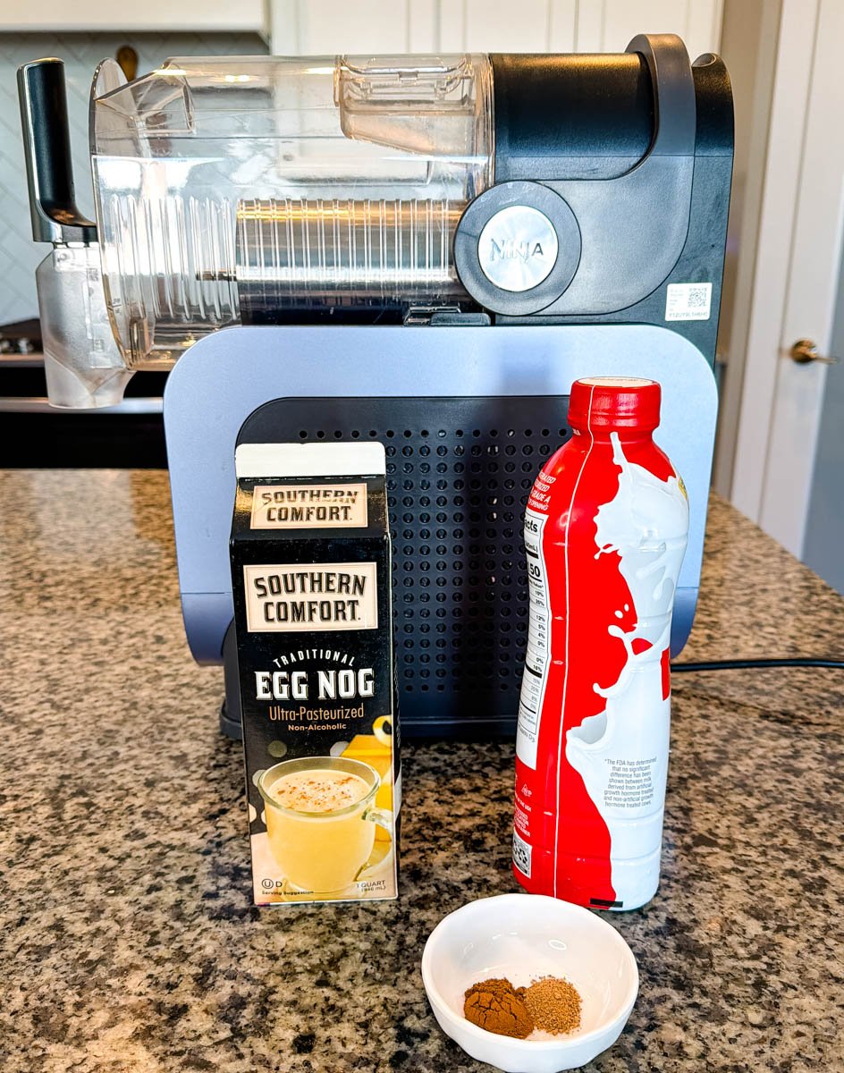 Ingredients for Ninja Slushi eggnog displayed in front of the machine, including Southern Comfort eggnog, whipped topping, and a small dish of cinnamon and nutmeg.