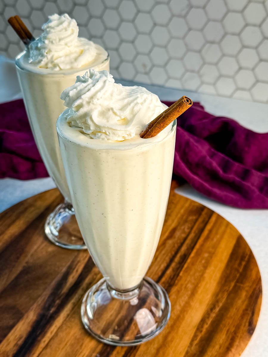 Overhead view of two glasses of frozen eggnog topped with whipped cream and cinnamon sticks, on a wooden board with a purple napkin in the background.