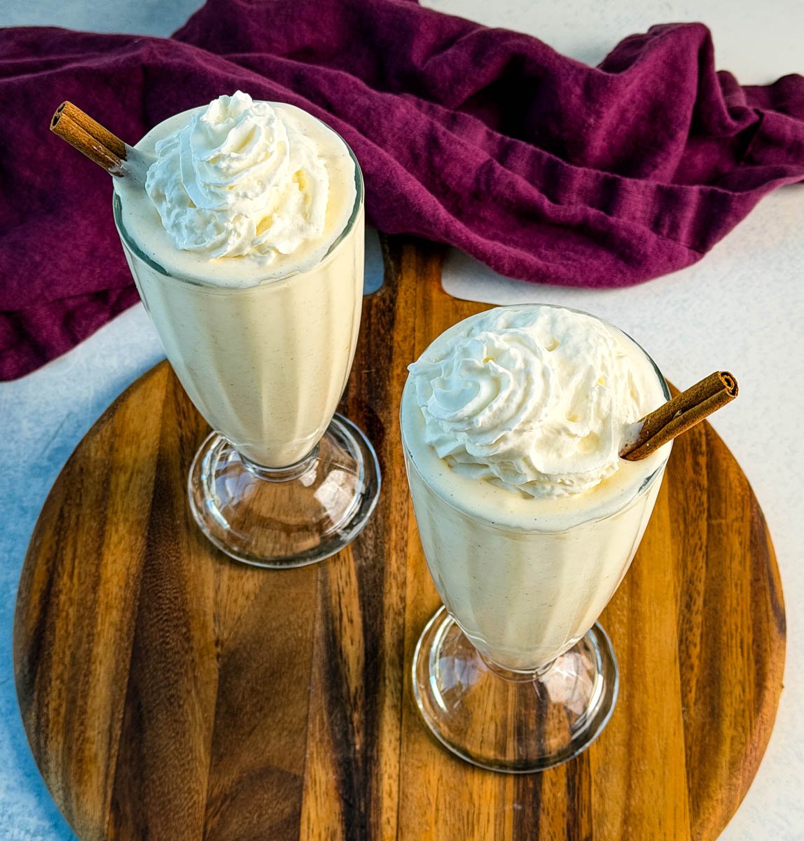 Overhead view of two glasses of frozen eggnog topped with whipped cream and cinnamon sticks, on a wooden board with a purple napkin in the background.