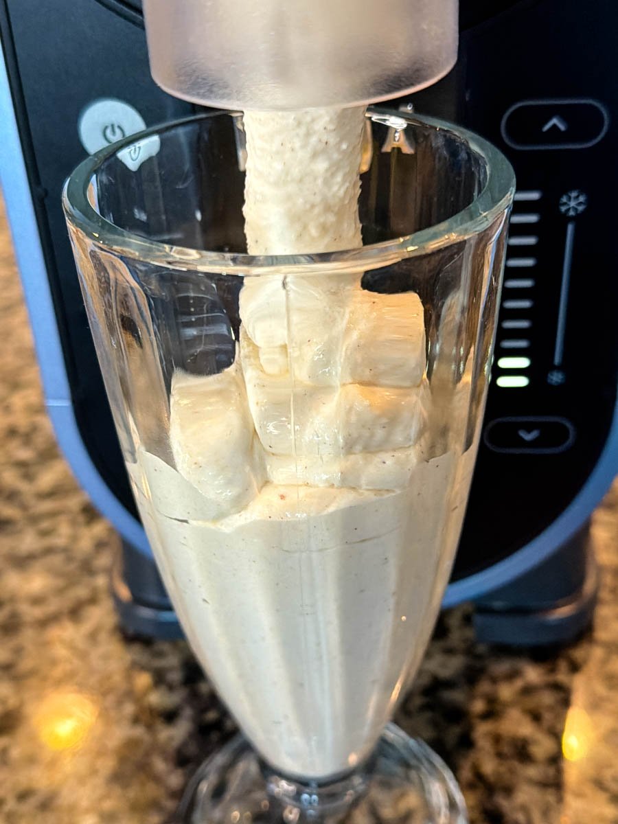 Frozen eggnog being dispensed from the Ninja Slushi machine into a tall glass, showing the smooth, creamy texture filling the cup.