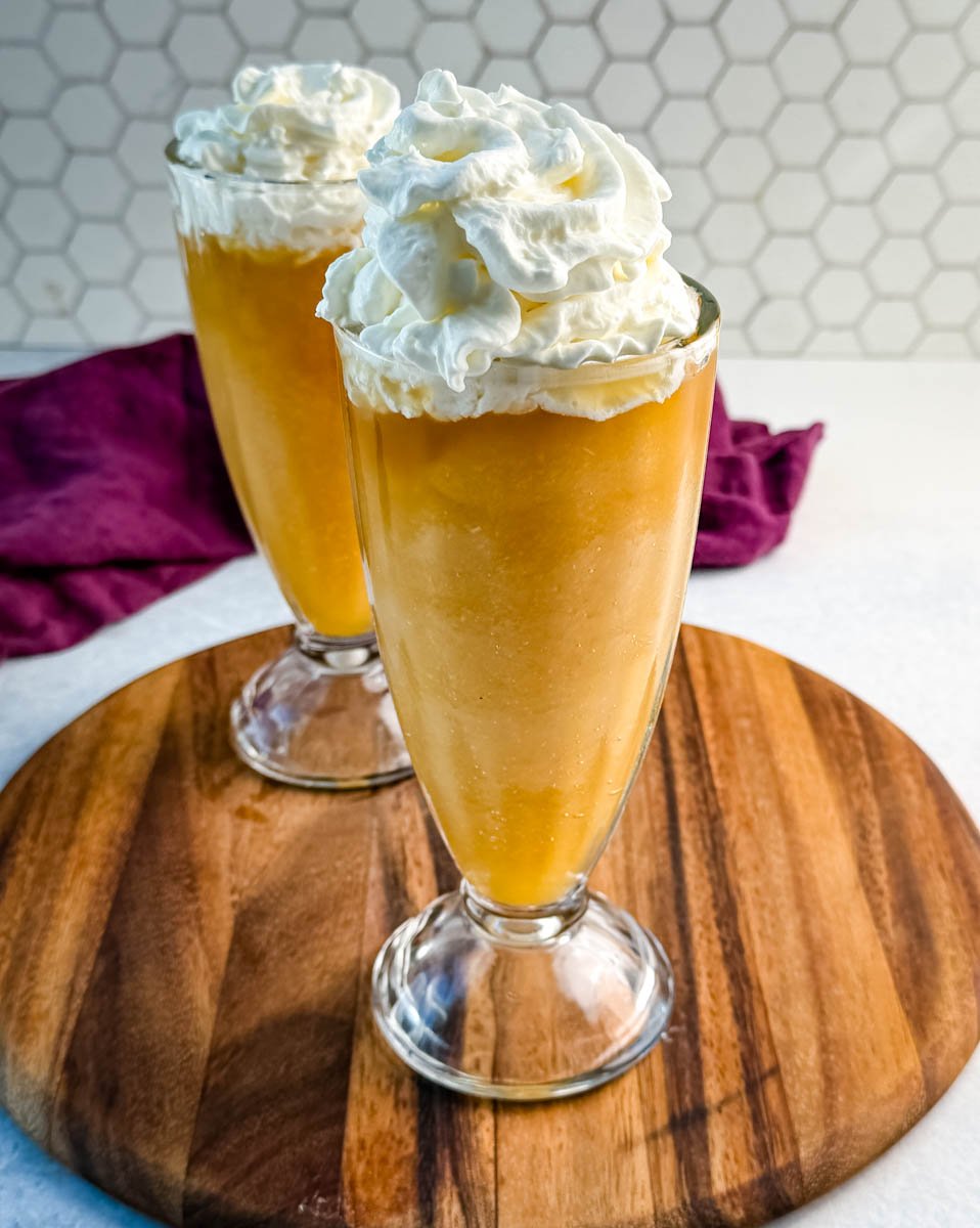 Two tall glasses filled with golden Butter Beer slushies, each topped with a large swirl of whipped cream, set on a wooden board with a hexagon-tile backsplash behind them.