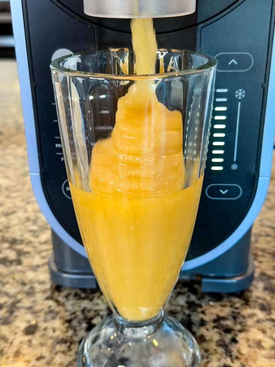 A thick, golden Butter Beer slushie pouring from a Ninja Slushi machine into a tall glass on a granite countertop.