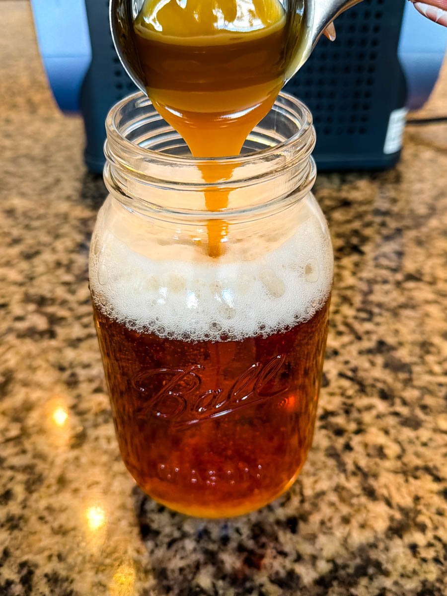 A spoonful of thick butterscotch sauce being drizzled into a mason jar filled with the golden Butter Beer mixture.