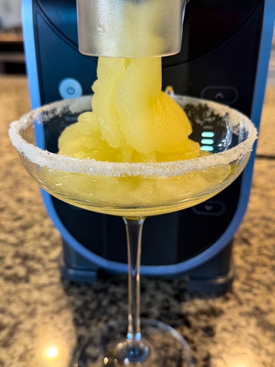 Frozen lemon drop slush dispensing from the Ninja slushi machine spout directly into a sugar-rimmed coupe glass, filling it with icy yellow slush.