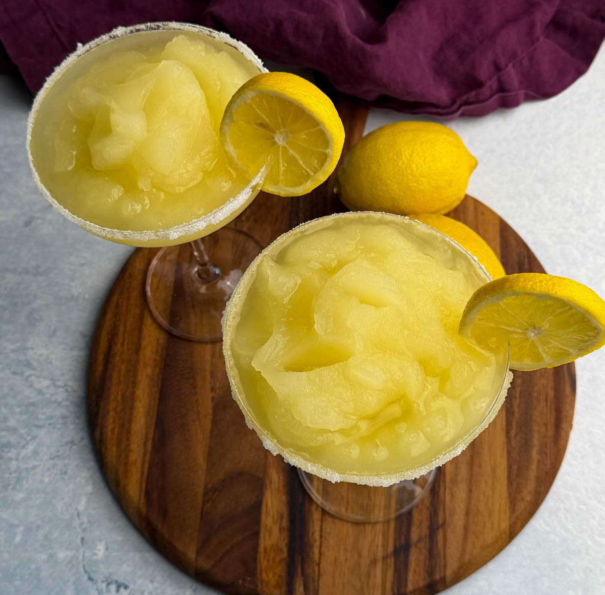 Overhead view of two coupe glasses filled with frozen lemon drop slush, each garnished with a lemon slice on the rim. Whole lemons sit beside the glasses on a round wooden board.