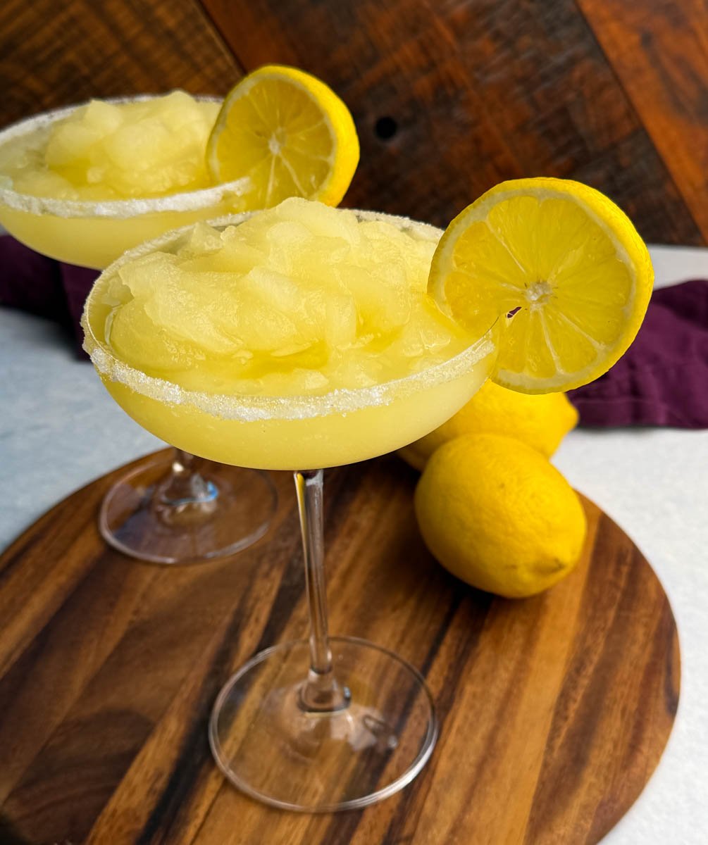 Close-up of a coupe glass filled with yellow frozen lemon drop slush, rimmed with sugar and garnished with a lemon slice, with another glass and whole lemons in the background.