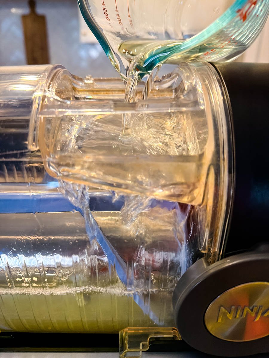 Liquid being poured from a glass measuring cup into the top of a Ninja slushi machine, filling the canister with the lemon mixture.