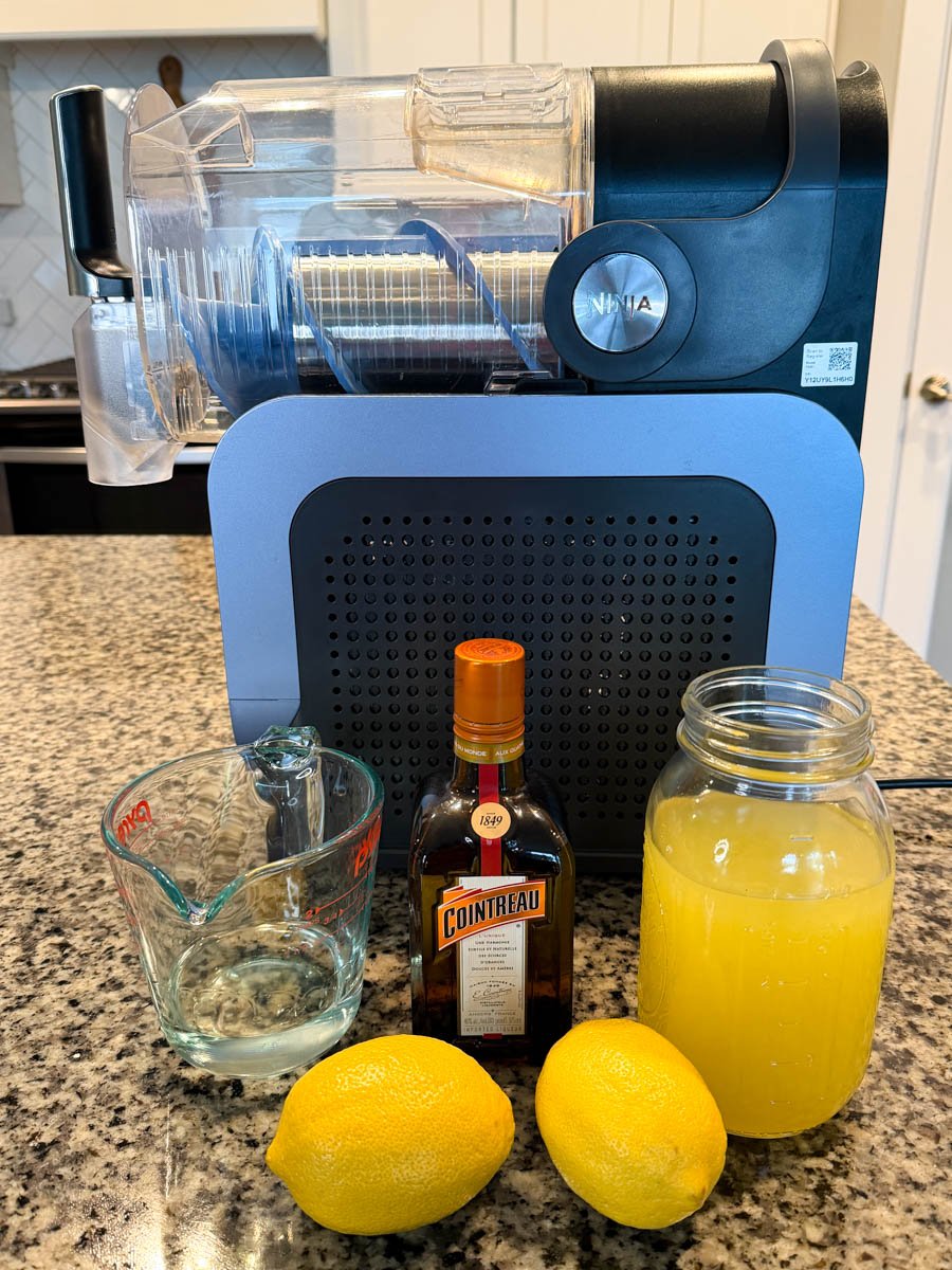 A Ninja slushi machine on a countertop with ingredients arranged in front: a measuring cup, a bottle of Cointreau, two whole lemons, and a large jar of lemon mixture.