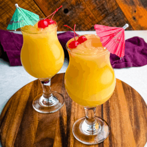Two frozen Mai Tai slushies in curvy hurricane glasses, each garnished with cherries and colorful umbrellas, placed on a round wooden board.