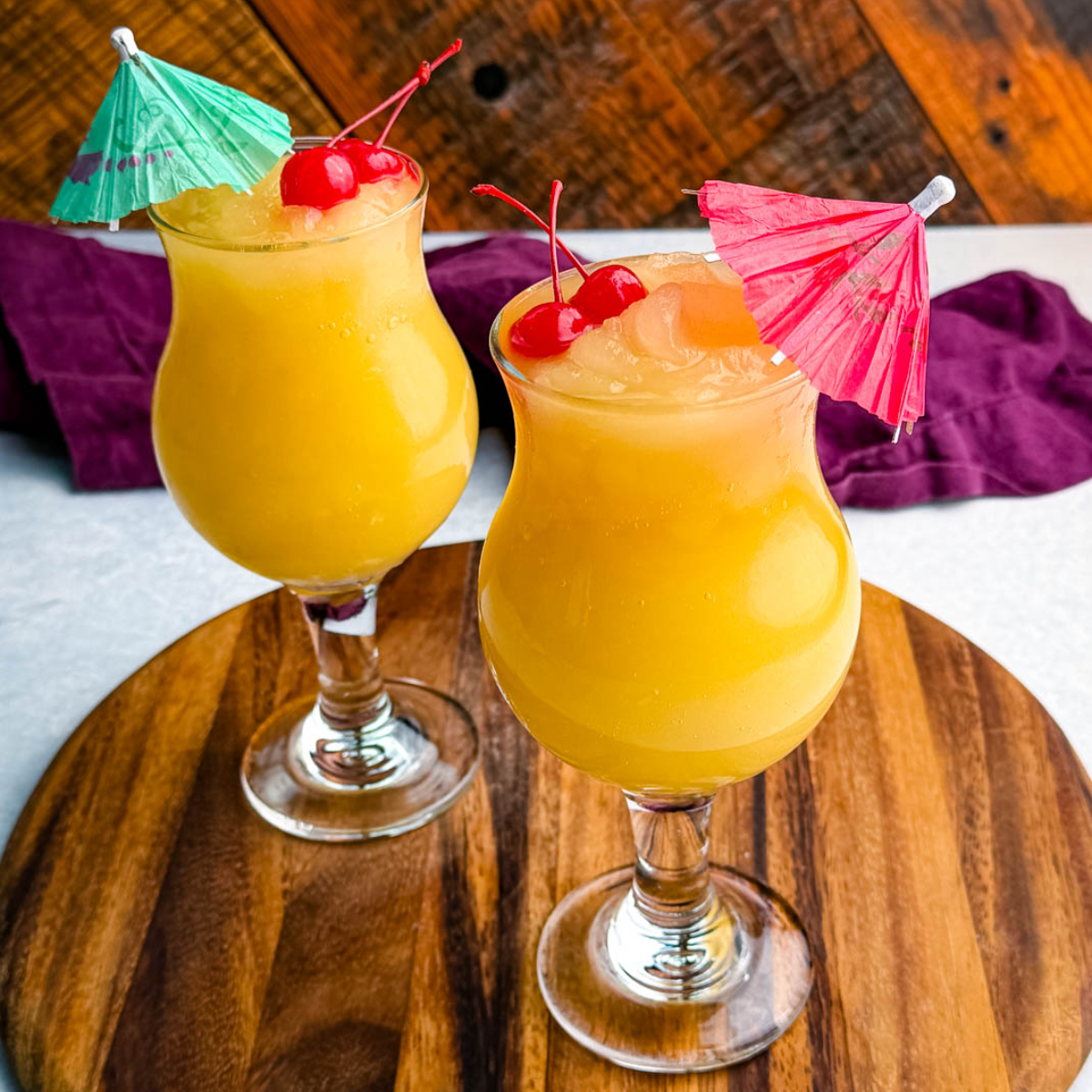 Two frozen Mai Tai slushies in curvy hurricane glasses, each garnished with cherries and colorful umbrellas, placed on a round wooden board.