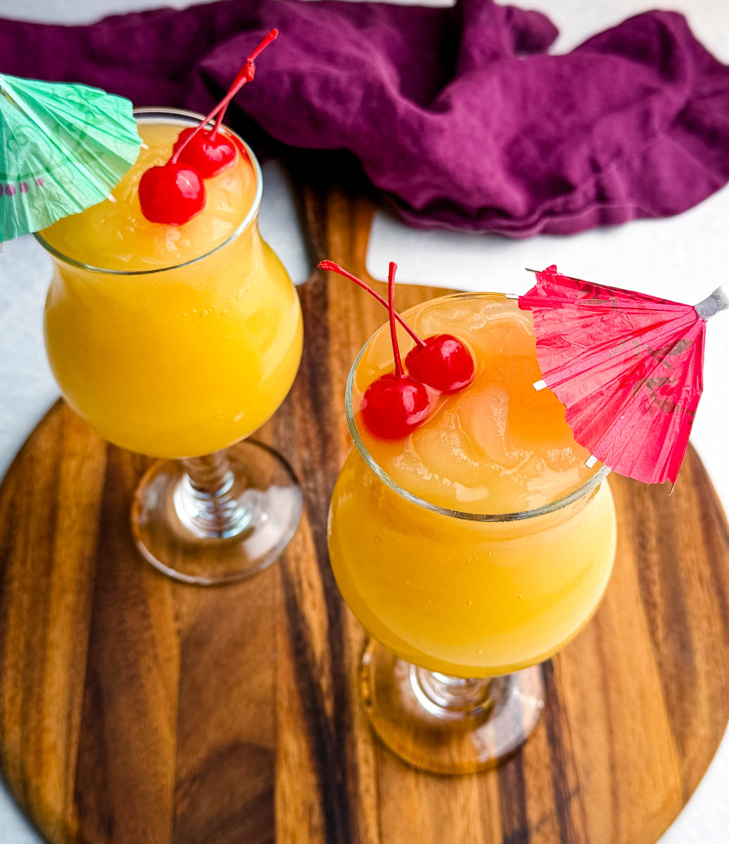 Two glasses of bright orange frozen Mai Tai slushies topped with maraschino cherries and paper drink umbrellas, sitting on a wooden board with a purple cloth in the background.