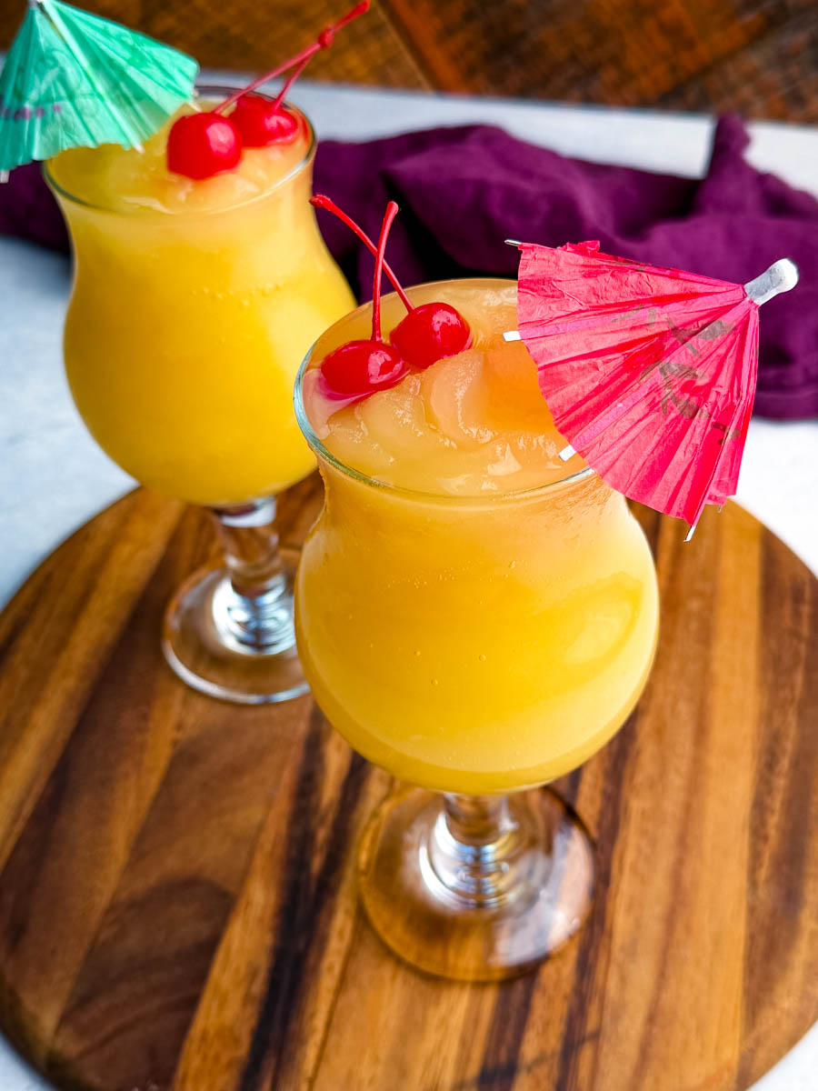 Two Mai Tai slushies garnished with maraschino cherries and paper umbrellas, displayed on a wooden board with a purple cloth behind them.
