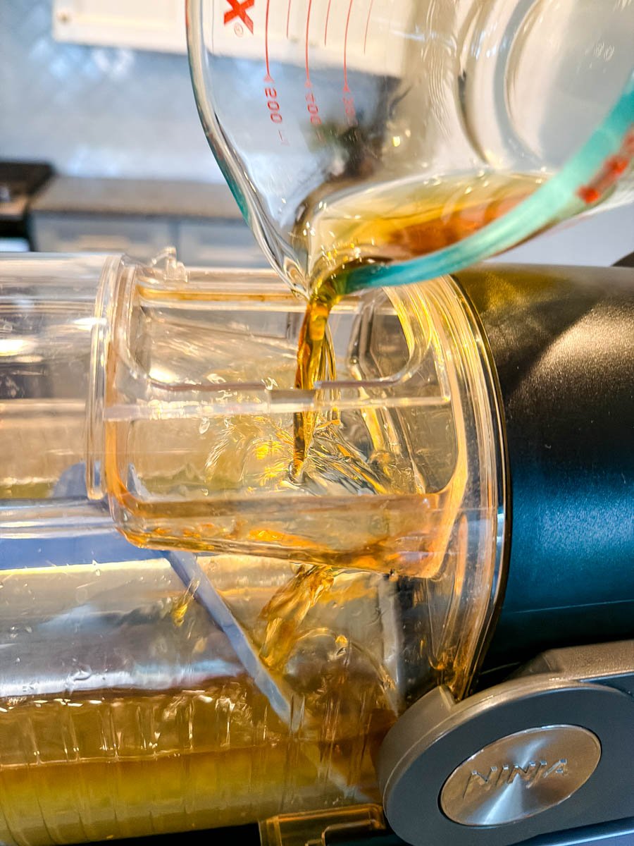 A measuring cup pouring a dark amber liquid—likely rum—into the top of the Ninja Slushi machine, with the mixture flowing into the canister.